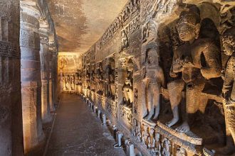 A long, narrow corridor carved into a rock face, lined with intricate sculptures and reliefs depicting various figures and scenes. Candles are placed along the floor, illuminating the space. This is likely a cave within the Ellora Caves complex in India.