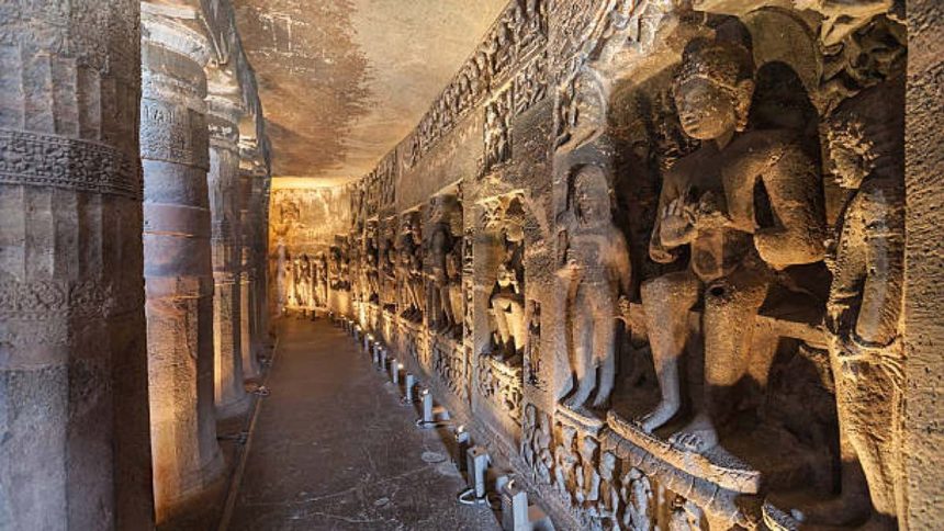 A long, narrow corridor carved into a rock face, lined with intricate sculptures and reliefs depicting various figures and scenes. Candles are placed along the floor, illuminating the space. This is likely a cave within the Ellora Caves complex in India.