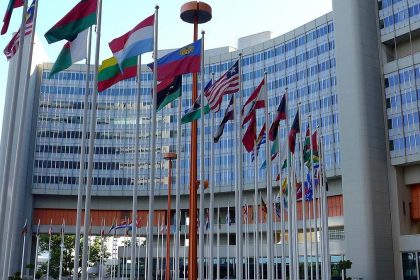 A large, modern building with a curved facade and numerous national flags flying from tall poles in front. The building appears to be the United Nations Headquarters.