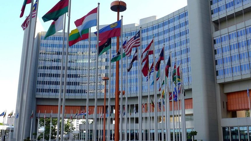 A large, modern building with a curved facade and numerous national flags flying from tall poles in front. The building appears to be the United Nations Headquarters.